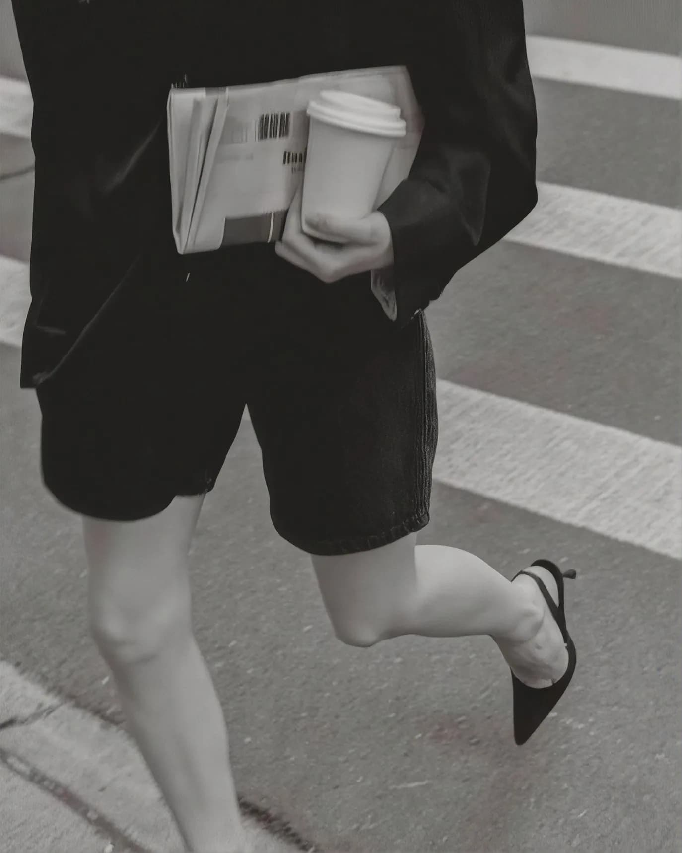 A woman crossing a zebra crossing in black shorts and slingback heels, a folded newspaper and a takeaway coffee under one arm. Black and white.